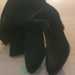 Svarta mockastövlar från Tamaris - Snygga svarta stövlar från Tamaris i mjuk mocka med spetsig tå och hög klack. Stövlarna har dragkedja på sidan för enkel påtagning och en stilren look som passar till många outfits.