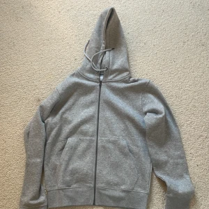 Grå hoodie från Jack & Jones - Snygg grå hoodie från Jack & Jones med dragkedja framtill och två stora fickor. Tröjan har huva med snörning och är tillverkad i mjuk bomullsmix. Säljer pga felköp och borttappat kvitto, aldrig använd. Se prislappen är ff kvar. Skicka 10/10
