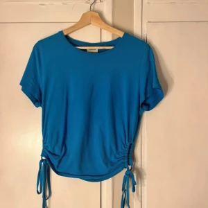 Blå topp med snörning från Noisy May - Säljer en blå topp från Noisy May i mjuk bomull. Modellen har korta ärmar, rund hals och snygg snörning med knyt i sidorna som ger en cool detalj. Perfekt för dig som gillar enkel men trendig stil.