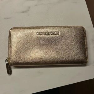 Guldig plånbok från Michael Kors - Snygg och stilren plånbok från Michael Kors i guldigt skinn med dragkedja runtom. Plånboken har flera kortfack, myntfack med dragkedja och logga i metall framtill. Perfekt för att hålla koll på alla dina kort och kontanter.