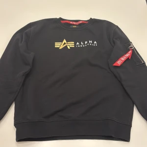 Svart Alpha Industries sweatshirt - Svart sweatshirt från Alpha Industries med guldigt logotryck på bröstet och vit text. Tröjan är gjort av ett lite tjockare material som värmer dig enkelt. Den har en cool design på vänster ärm med en rödfärgad dragkedja. Den är använd runt 2-3 gånger alltså den är som en helt ny. Storleken är M och kan passa folk som har storleken L. Pris kan diskuteras.