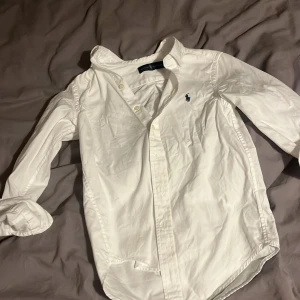 Vit skjorta från Ralph Lauren - Vit skjorta från Ralph Lauren med klassisk krage och knappar framtill. Skjortan har lång ärm och den ikoniska broderade Polo-loggan på bröstet. Tillverkad i mjuk bomull för en clean och stilren look. Perfekt för dig som gillar tidlös design och snygga detaljer.