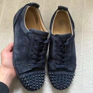 Christian Louboutin svarta sneakers med nitar - Säljer ett par svarta sneakers från Christian Louboutin i mocka med ikoniska röda sulor. Skorna har snörning och coola svarta nitar på tån för en edgy vibe. Perfekta för dig som vill sticka ut med lyxiga detaljer och streetstyle-känsla.
