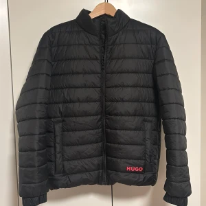 Svart pufferjacka från HUGO - Svart pufferjacka från HUGO med röd logga på framsidan. Jackan har quiltad design, ståkrage och dragkedja framtill. Två stora fickor på sidorna och en innerficka med dragkedja. Perfekt för kalla dagar och har en clean, modern look. Orginal pris 3.100kr