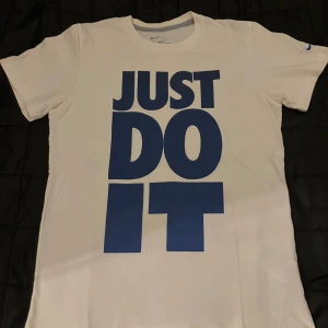 Vit Nike t-shirt med blå tryck - Nike t-shirt i vitt med stort blått 'JUST DO IT'-tryck på framsidan. Slim fit-modell med korta ärmar och rund halsringning. Klassisk och sportig stil, perfekt för dig som gillar streetwear och sportmode.