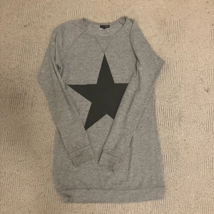 Grå långärmad tröja med svart stjärna - Grå långärmad tröja från KappAhl med en stor svart stjärna på bröstet. Mjuk och skön i bomullsmix, perfekt för chill dagar. Enkel rund halsringning och rak passform. Snygg att matcha med jeans eller leggings.