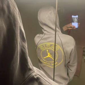 Grå Jordan PSG hoodie med gul logga - Säljer en grå hoodie från Jordan x Paris Saint-Germain med gul Jumpman-logga och PSG-märke på bröstet samt stor gul logga på ryggen. Tröjan har huva med snörning och en stor magficka. Perfekt för dig som gillar streetwear och sportig stil.