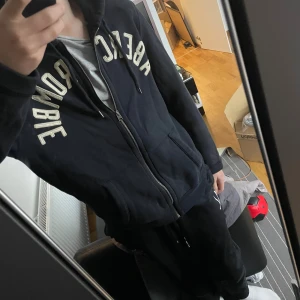 Svart hoodie från Abercrombie & Fitch - Svart hoodie från Abercrombie & Fitch med broderad logga i vitt över bröstet. Tröjan har huva med snörning, dragkedja framtill och två stora fickor. Tillverkad i mjuk bomullsblandning, perfekt för chill dagar. Klassisk och stilren look.