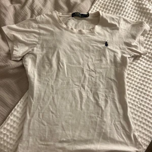 Vit t-shirt från Polo Ralph Lauren - Vit t-shirt från Polo Ralph Lauren med klassisk rund hals och liten mörkblå broderad logga på bröstet. Gjord i mjuk bomull och har en enkel, clean look som passar till allt. Perfekt basic-plagg för garderoben.