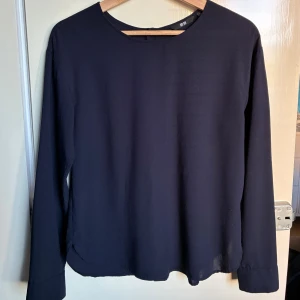 Mörkblå långärmad blus från Uniqlo - Snygg mörkblå blus från Uniqlo i viskos och polyester. Rak modell med rund halsringning och knappar längs ryggen som cool detalj. Perfekt till jeans eller kjol för en clean look. Långärmad och enkel att styla till olika outfits.
