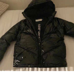 Svart pufferjacka från Calvin Klein Jeans - Snygg svart pufferjacka från Calvin Klein Jeans med glansig finish och huva. Jackan har dragkedja framtill och diskret logga på insidan. Perfekt för kalla dagar och ger en cool streetstyle-vibe.