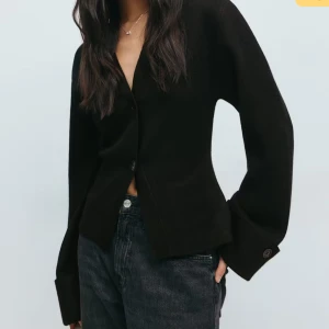 Svart kofta Gina  - Svart kofta med v-ringning från Gina Tricot. Modellen har långa ärmar med breda manschetter och knappar, samt två knappar framtill. Koftan är rak i modellen och har en stilren, minimalistisk look som passar perfekt till jeans. Endast använd en gång så mycket bra skick🥰