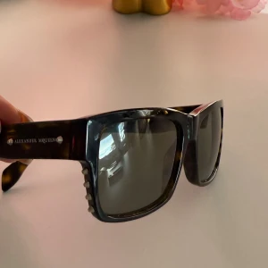 Svarta solglasögon Alexander McQueen - Snygga solglasögon från Alexander McQueen med bred, fyrkantig båge i svart och mörkbrun sköldpaddsmönstrad plast. Glasen är mörka och sidorna har coola nitar som detalj. Perfekt accessoar för att lyfta din stil.