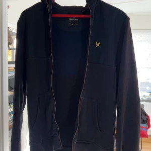 Svart hoodie från Lyle & Scott med dragkedja - Svart hoodie från Lyle & Scott med gul broderad logga på bröstet. Tröjan har huva, hel dragkedja och två fickor framtill. Tillverkad i mjuk bomullsblandning, perfekt för chill dagar. Klassisk och stilren design med lång ärm.