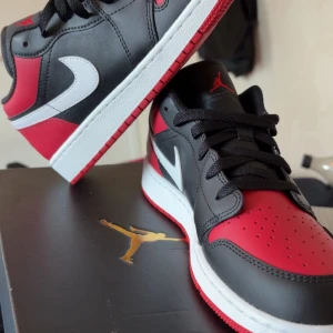 Nike Air Jordan 1 Low svart/röd/vit - Säljer ett par Nike Air Jordan 1 Low i svart, rött och vitt. Klassisk sneaker med vit swoosh, svart snörning och röd yttersula. Skorna har rund tå, platt sula och är tillverkade i skinn. Perfekt för dig som gillar streetstyle och ikoniska sneakers.har aldrig använt dom heller 