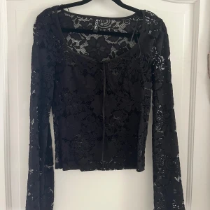 Svart spetsblus från Pieces - Svart blus från Pieces i storlek S, i genombruten spets med blommigt mönster. Modellen har långa ärmar och vida ärmar, en rundad halsringning med knyt framtill. 