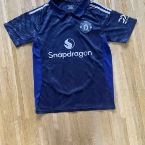 Manchester United blå fotbollströja - Snygg blå Manchester United fotbollströja med Snapdragon-logga på bröstet och klubbmärke. Tröjan har korta ärmar, krage och diskret mönster i tyget. Materialet är lätt och andas, perfekt för träning eller match. Silverfärgade detaljer på axlarna och tryck på ärmen.