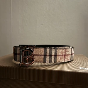 Burberry vändbart bälte med monogram - Snyggt vändbart bälte från Burberry med klassiskt rutigt mönster i beige, svart och rött på ena sidan och svart präglad design på andra. Stort metallspänne med Burberrys ikoniska TB-monogram. Perfekt accessoar för att lyfta din outfit.