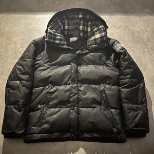 Svart pufferjacka från Burberry - Snygg svart pufferjacka från Burberry med huva och rutigt innerfoder. Jackan har dragkedja, knappar och två fickor med dragkedja framtill. Stor Burberry-patch på ärmen ger en exklusiv vibe. Perfekt för kalla dagar när du vill hålla stilen. Hör av vid minsta fundering 💫