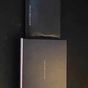 Svart plånbok från Tommy Hilfiger - Snygg svart plånbok från Tommy Hilfiger i skinn med stilrena ränder i marinblått, vinrött och vitt på insidan. Perfekt storlek för kort och sedlar, med klassisk logga på framsidan. Kommer med originalask.