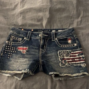 Miss Me denimshorts med nitar och patch - Snygga blå jeansshorts från Miss Me med slitna kanter, nitar och coola patchdetaljer i rött, vitt och silver. Shortsen har låg midja, klassiska fickor och broderade mönster med amerikansk flaggstil. Perfekt för dig som gillar edgy och unik streetstyle.