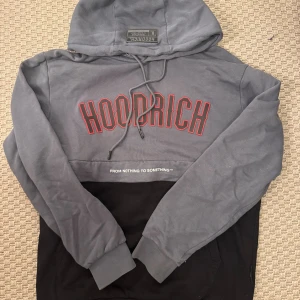 Grå och svart hoodie från Hoodrich - Säljer en snygg tvåfärgad hoodie från Hoodrich i grått och svart med röd logga framtill. Tröjan har huva med snörning, muddar vid ärmslut och midja samt tryck både fram och bak. Perfekt för streetwear-stil och chill dagar.