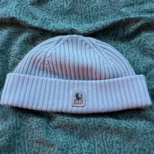 Ljusgrå ribbad mössa Parajumpers - Snygg ljusgrå ribbstickad mössa från Parajumpers med uppvikt kant och klassisk PJS-logga framtill. Perfekt för kalla dagar och enkel att matcha med din jacka. Materialet är mjukt och värmande, och designen är clean med en modern vibe.