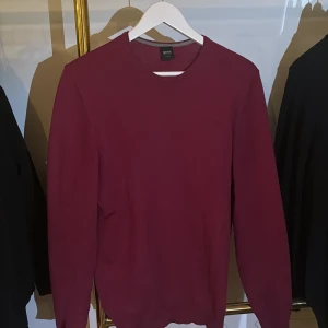 Vinröd ulltröja från Hugo Boss - Vinröd stickad tröja från Hugo Boss i slim fit-modell. Tillverkad i 100% ull med rund halsringning och långa ärmar. Perfekt för dig som gillar stilrena och klassiska plagg med en modern passform.