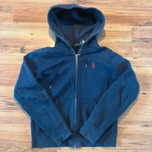 Marinblå hoodie från Polo Ralph Lauren - Snygg marinblå hoodie från Polo Ralph Lauren med dragkedja och huva. Tröjan har det klassiska röda broderade logotypen på bröstet och är tillverkad i mjuk bomull. Perfekt för chill dagar eller när du vill ha en stilren streetlook.