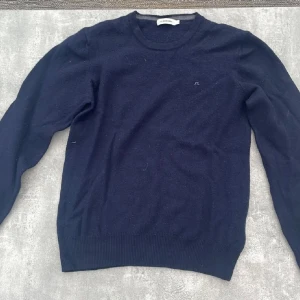 Mörkblå kashmirtröja J.Lindeberg - Mörkblå stickad tröja från J.Lindeberg i mjuk kashmir. Klassisk rund halsringning, diskret logga på bröstet och ribbade muddar vid ärmslut och nederkant. Perfekt för lager-på-lager och stilren look.