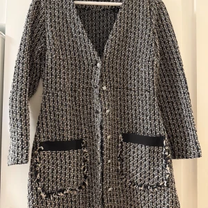 Svartvit rutig kappa från Zara - Stilren svartvit kappa från Zara i rutigt mönster med grovstickad känsla. Långärmad modell med v-ringning, dekorativa knappar och två stora fickor framtill med fransade kanter och svarta band. Perfekt för dig som gillar klassisk men trendig look.