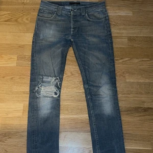 Blå slitna jeans från Nudie Jeans - Snygga blå jeans från Nudie Jeans med coolt slitet och trasigt parti på vänster knä. Klassisk femficksmodell med raka ben och normal passform. Jeansen har en tvättad look och är tillverkade i robust denim.