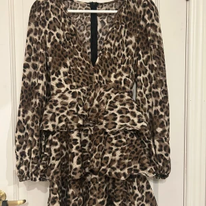 Leopardmönstrad volangklänning - Kort klänning med leopardmönster och volanger. Djup v-ringning, långa ärmar med resår vid handlederna och dragkedja i ryggen. 