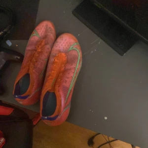Nike Mercurial Vapor fotbollsskor orange - Säljer ett par Nike Mercurial Vapor fotbollsskor i orange med gröna detaljer och snörning. Skorna har en tunn syntetisk ovandel med ACC-teknologi och mönstrad sula med dobbar i lila och orange för grymt grepp på planen. Perfekta för dig som vill sticka ut på matchen!