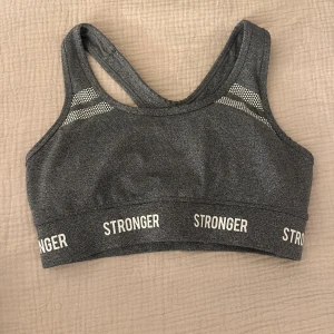 Grå sporttopp från Stronger - Säljer en grå sporttopp från Stronger med brottarrygg och vita logotyper runt resåren. Toppen har svarta meshpaneler på insidan och diskreta mönsterdetaljer vid axlarna. Perfekt för dig som vill ha en snygg och bekväm topp till träningen.