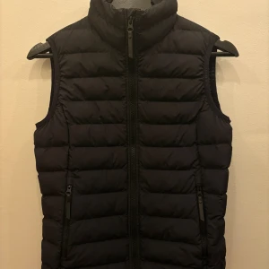 Svart dunväst Peak Performance XS - Svart dunväst från Peak Performance i storlek XS. Västen har quiltad design, hög krage och två fickor med dragkedja framtill. Perfekt för lager-på-lager och har en stilren look med diskret logga på bröstet.