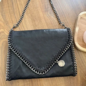 Stella McCartney Svart axelväska - Snygg svart axelväska från Stella McCartney med kedjedetaljer runt kanterna och på axelremmen. Väskan har ett kuvertformat lock, dekorativ medaljong och dragkedja. Insidan är fodrad i orange satin med logotypmönster. Perfekt för dig som gillar statement-accessoarer.