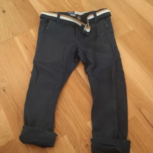 Mörkblå chinos från Okaidi med bälte - Snygga mörkblå chinos från Okaidi i slim fit-modell. Byxorna har uppvikta benslut, sidofickor och bakfickor samt ett randigt tygbälte i beige, vitt och svart. Tillverkade i bomull för en skön känsla och stilren look.
