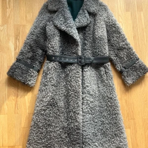 Kappa i lammskinn - Fantastiskt vacker och exklusiv kappa i äkta gotländskt lammskinn, känd för sin mjukhet, glans och naturliga lock. Ett hantverksmässigt plagg som kombinerar klassisk stil med kyliga vinterdagar – lika fin till vardags som till mer eleganta tillfällen. Kappan har en skimrande insida som skiftar mellan gröna och lila toner.  Längd (nacke till nederdel): 115 cm •	Ärmlängd: 50 cm •	Ärmhåla till nederdel: 80 cm •	Färg: Naturlig grå/silvernyans med vacker lyster •	Ny skick – välvårdad, utan skador.