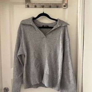 Grå stickad tröja från Gina Tricot - Mysig grå stickad tröja från Gina Tricot med krage och v-ringning. Tröjan har långa ärmar och breda ribbade muddar vid ärmslut och nederkant. Perfekt för lager-på-lager och chill dagar.