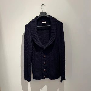 Mörkblå stickad kofta från Topman - Mysig mörkblå stickad kofta från Topman med grov kabelstickning och stora bruna knappar. Koftan har en bred sjalkrage och långa ärmar, perfekt för kyliga dagar. Snygg och klassisk modell som funkar till det mesta. Den saknar en knapp men inget man märker vid användning, hör av er vid frågor!