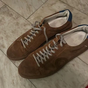 Bruna skor Oliver Cabell - Säljer ett par bruna sneakers i mocka med rund tå. Skorna har en diskret cirkeldetalj på sidan och blå detalj vid hälen. Snygg och enkel design som passar till det mesta.