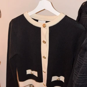 Svart kofta med gulddetaljer från H&M - Stilren svart kofta från H&M med kontrasterande vita kanter och dekorativa guldfärgade knappar. Koftan har två små fickor framtill med vita detaljer och är långärmad. Perfekt för att lyfta din outfit med en klassisk touch.