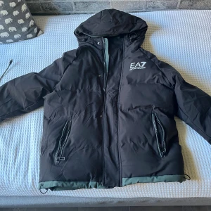 Svart pufferjacka EA7 Emporio Armani - Svart pufferjacka från EA7 Emporio Armani med huva och stora logotexter både fram och bak. Jackan har dragkedjor på fickorna, ribbade muddar och grå detaljer längs nederkant och ärmar. Perfekt för dig som vill ha en stilren och varm jacka med streetkänsla.