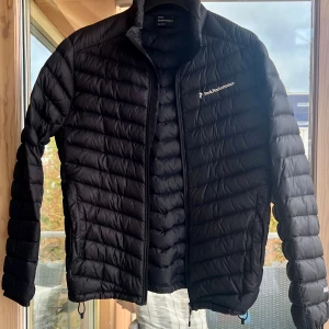 Svart dunjacka från Peak Performance - Svart, tunn dunjacka från Peak Performance med stilren design och diskret logga på bröstet. Jackan har horisontella quiltade paneler, dragkedja framtill och hög krage. Perfekt för kyliga dagar och enkel att matcha med din vardagsstil.