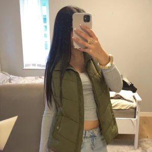 Olivgrön quiltad dunväst från H&M - Snygg olivgrön dunväst från H&M med quiltad design och hög krage. Västen har dragkedja framtill och tryckknappar vid fickorna. Perfekt att slänga över en hoodie eller långärmad tröja för en chill look.