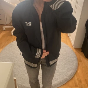 Svart EA7 bomberjacka med ränder - Säljer en svart jacka  från EA7 Emporio Armani med vita ränder på muddar och nederkant. Jackan har dragkedja framtill och logga på bröstet. Materialet är polyester och jackan har en klassisk, normal passform. Perfekt för dig som gillar stilrena streetwear-vibbar. Den är använd max 5 gånger så som helt ny i stort sätt. Det är storlek xs men den är stor i storleken. Köpt i affär över 1000