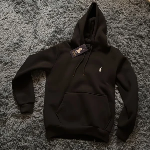 Svart hoodie från Polo Ralph Lauren - Svart hoodie från Polo Ralph Lauren med klassisk vit broderad logga på bröstet. Tröjan har huva med snörning, känguruficka och ribbade muddar. Perfekt för en chill och stilren look.obs inte original, men fortfarande väldigt skön ull insida