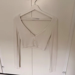 Vit croppad ribbad topp från Bershka - Säljer en vit croppad topp från Bershka med lång ärm och v-ringning. Toppen är ribbad och har två små knappar framtill. Perfekt för dig som gillar stilrena och enkla plagg med snygg passform.