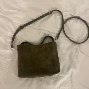 Grön mocka axelremsväska - Snygg olivgrön axelväska i mjuk mocka med justerbar rem och silverfärgade detaljer. Väskan har en praktisk innerficka med dragkedja och magnetstängning. Nypris var 600 tror jag och den är nästan aldrig använd.